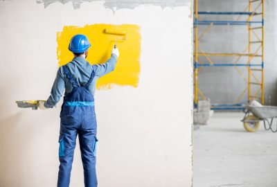 Workman in uniform painting wall with yellow paint at the construction site indoors